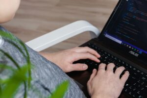 Person typing code on an ASUS laptop while sitting in a white chair, with blurred green plant leaves in the foreground.